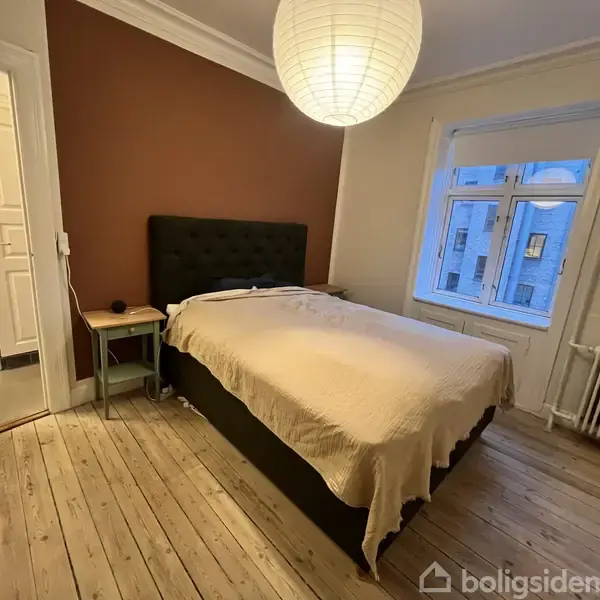En seng med beige sengetæppe står i et soveværelse med trægulv. En papirlampe hænger over sengen, og der er en brun væg bagved. En dør fører til en anden del af lejligheden.