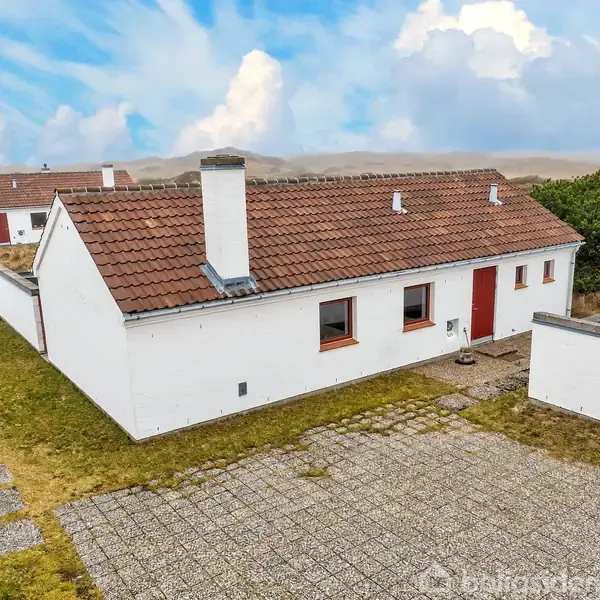 Hvidt murstenshus med rødt tag står stille på en flisebelagt gårdsplads omgivet af græsarealer. Andre lignende huse ses i baggrunden under en blå, skyet himmel.