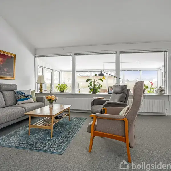 En stue med grå sofaer og en lænestol omkranser et træbord med blomster. Store vinduer i baggrunden giver udsigt til udendørs lys. Vægkunst hænger på venstre væg.