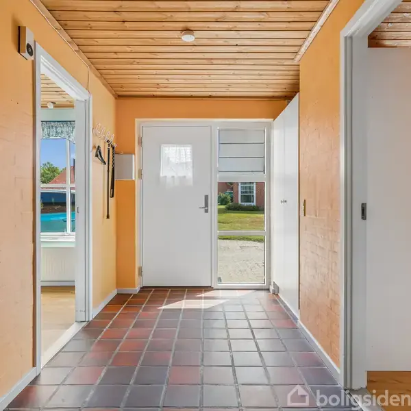 En entré med sort-røde fliser og orange vægge fører til en glasdør, der åbner ud mod en have. To åbne døråbninger viser tilstødende rum med farvede vægge.