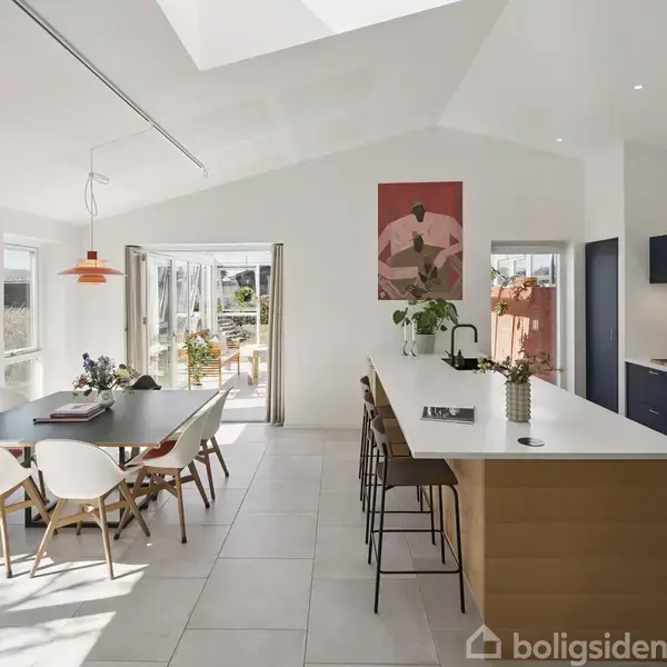 Et moderne køkken-alrum med lysindfald, hvor et spisebord er omgivet af stole til venstre, mens en stor køkkenø med barstole ses til højre. Dobbeltdøre åbner mod en terrasse.