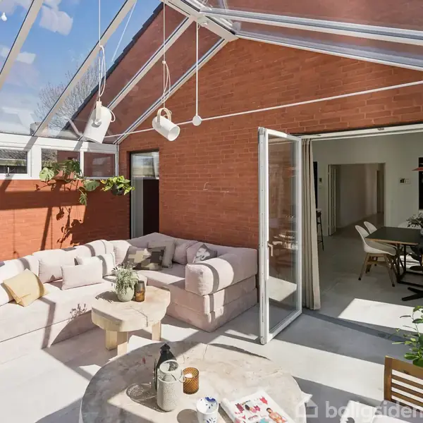 Glasoverdækket terrasse med beige hjørnesofa og sofabord. Terrassen er tilstødende en indendørs spisestue, synlig gennem åbne dobbeltdøre. Murstensvægge og planter skaber en hyggelig atmosfære.