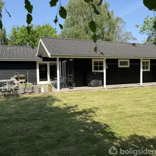 Sort træhus står stille i en grøn have med græsplæne. En terrasse med havemøbler er til venstre, omgivet af træer og klare, blå himmel.
