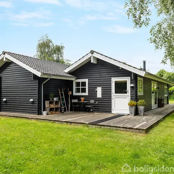 Et sort, træbeklædt hus står på en grøn græsplæne. En træterrasse omkranser huset, og omgivelserne inkluderer træer og buske under en blå himmel.