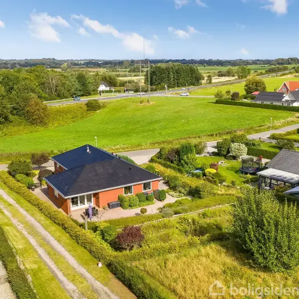 Rødt murstenshus med sort tag står i centrum, omgivet af en velplejet have med græsplæne og buske. Omgivende landbrugsarealer og enkeltstående huse ses i baggrunden under en blå himmel.