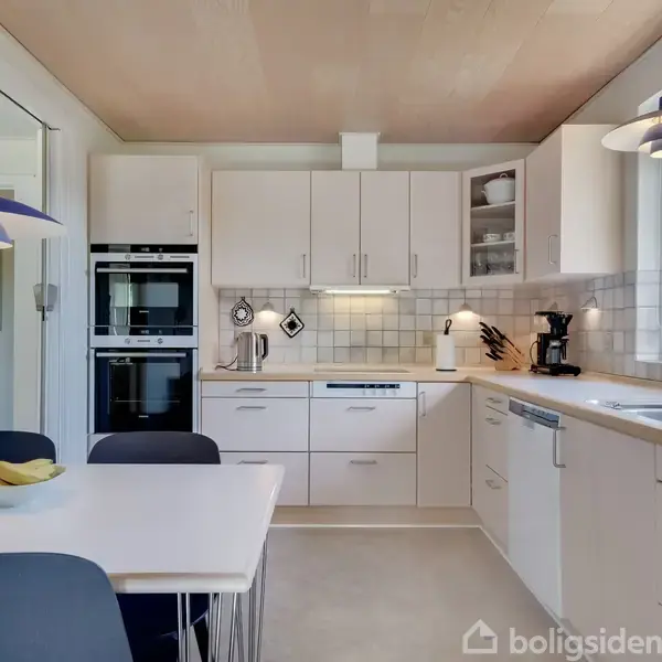 Et lyst køkken med hvide skabe; en kaffemaskine og en blender står på bordet. To blå-hvide lamper hænger over spisebordet med en skål bananer.