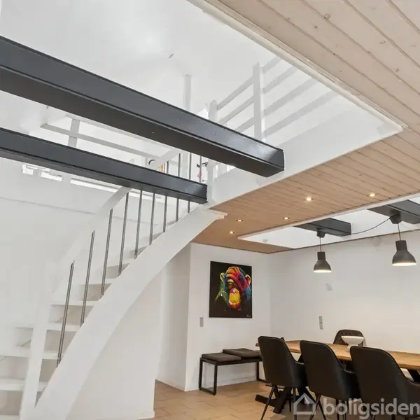 Rund trappe strækker sig opad mod en øverste etage i et moderne rum. Ved siden af står et spisebord med seks stole, oplyst af loftslamper. Kunst hænger på væggen.