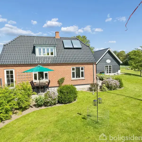 Et rødt murstenshus med sort tag står i en velplejet have. En terrasse med parasol ses foran huset. Græsplænen omgiver ejendommen med spredte buske og træer.