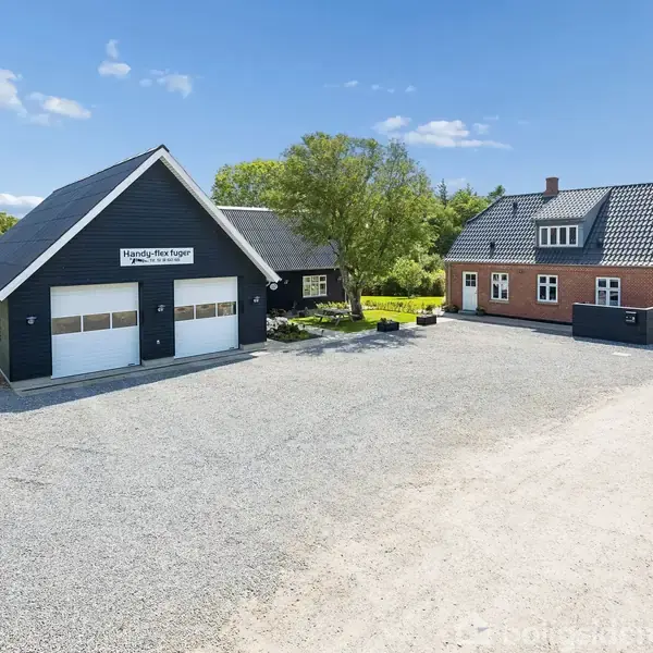 Murstenshus og garagebygning står side om side på en stor grusplads omgivet af grønne områder. Skilt på garagen: "Handels-Post-Rogn v/ Jørgen Kristensen."