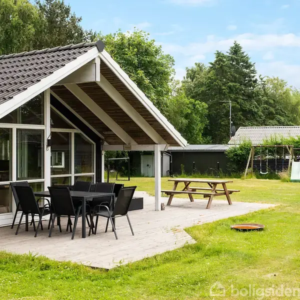 En modernistisk sommerhytte med møbleret terrasse står i en velholdt have. Hytten har store vinduespartier under et skråtag. Græsplænen omkranser et gyngestativ og piknikbord.