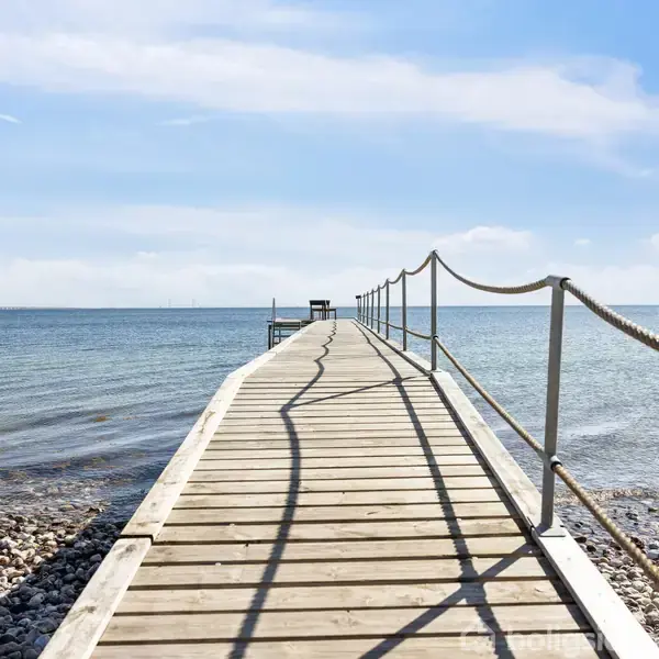 En lang træbro strækker sig ud i havet, omringet af klart blåt vand og en stenet kystlinje under en skyfri, solrig himmel.