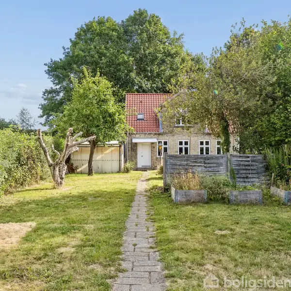 Et lille hus med rødt tag og hvide vinduer står i en have. En flisebelagt sti fører gennem græsplænen mod huset, omgivet af træer og buske.