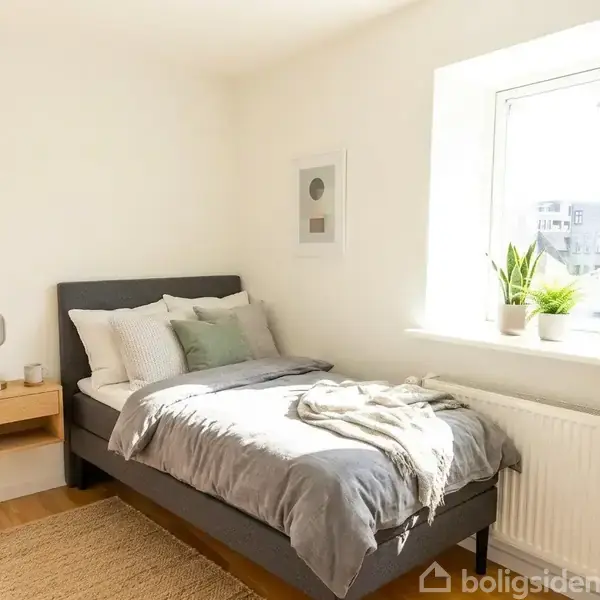 Seng med gråt sengetøj står ved siden af et vindue, med en plante på vindueskarmen. Et natbord med lampe hænger på væggen. Rummet er lyst og minimalistisk.