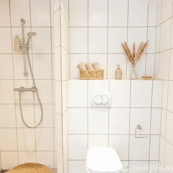 Bruseområde med brusehoved, flisebeklædte vægge. Til højre, et toilet, med kurv indeholdende sammenrullede håndklæder og dekorationer på hylde. Kurv med tørrede blomster i lys miljø.
