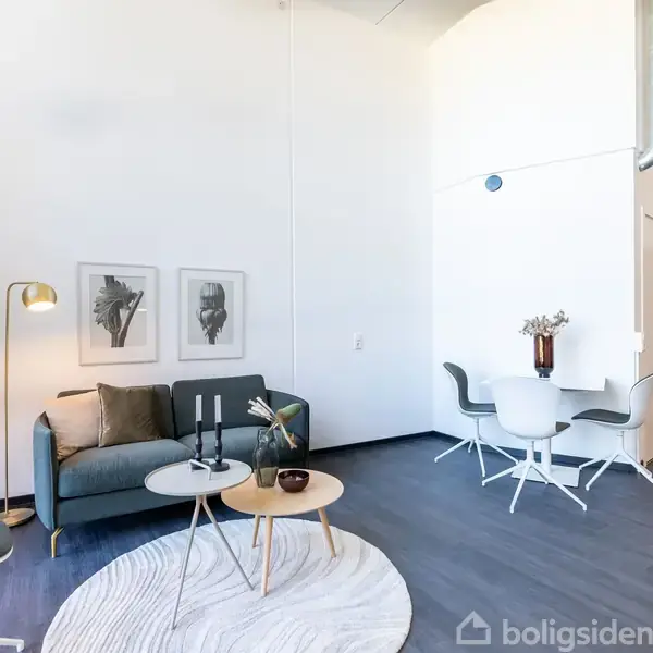 Stue med sofa og bordlampemann pynter op ved siden af moderne stole og et rundt bord i et lyst rum. Væggen har to kunstværker, og gulvet er mørkt.