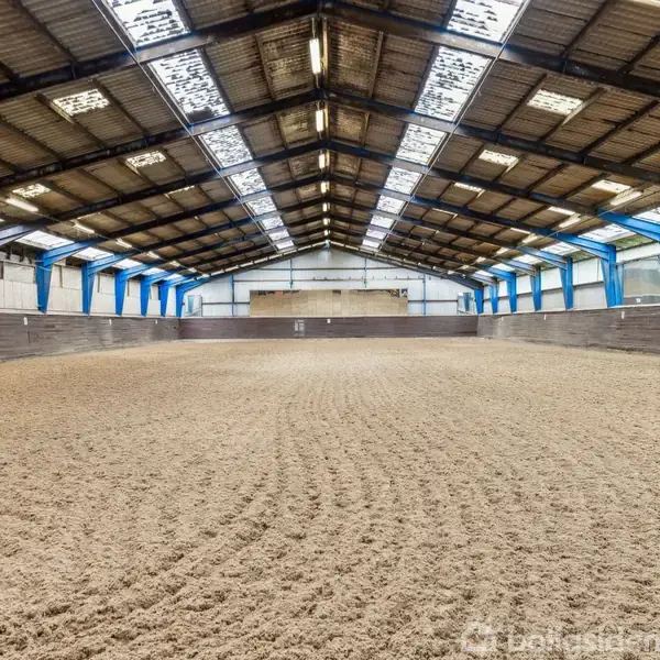 Ridebane med sandgrund, ingen aktivitet, beliggende i en stor indendørs arena med højt loft og metalbjælker, oplyst af ovenlys og flankerede blå supportstrukturer.