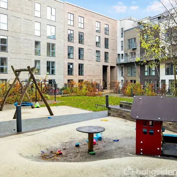 Legeplads med gynger og legehus står stille i en gårdhave omgivet af moderne lejlighedsbygninger, med farverige efterårsblade og klare himmel i baggrunden.