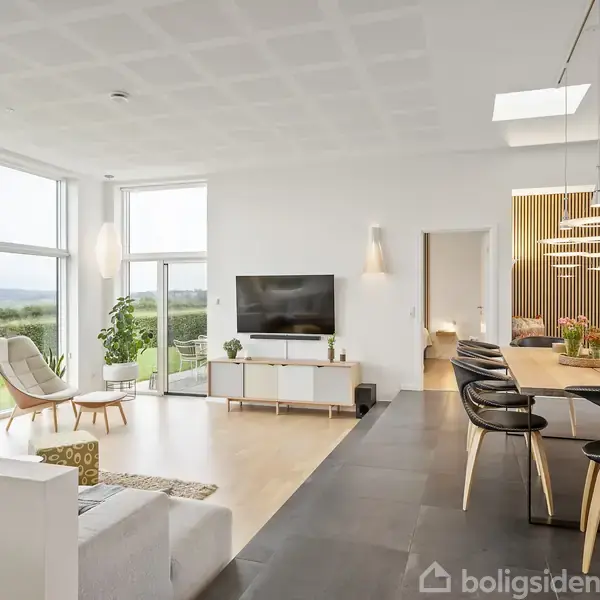 En moderne stue med sofa, stole og planter ved store vinduer. Et spisebord står til højre med stole og lamper over. Der er en tv-skærm. Udsigt til natur udenfor.
