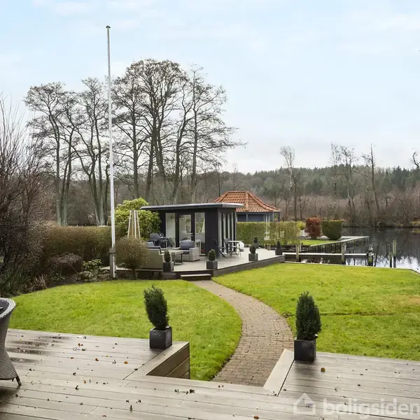 Terrasse med træbord og stole vender ud mod græsplæne og sø. En sti fører til et lysthus ved vandet, omgivet af buske og træer i en park-lignende have.