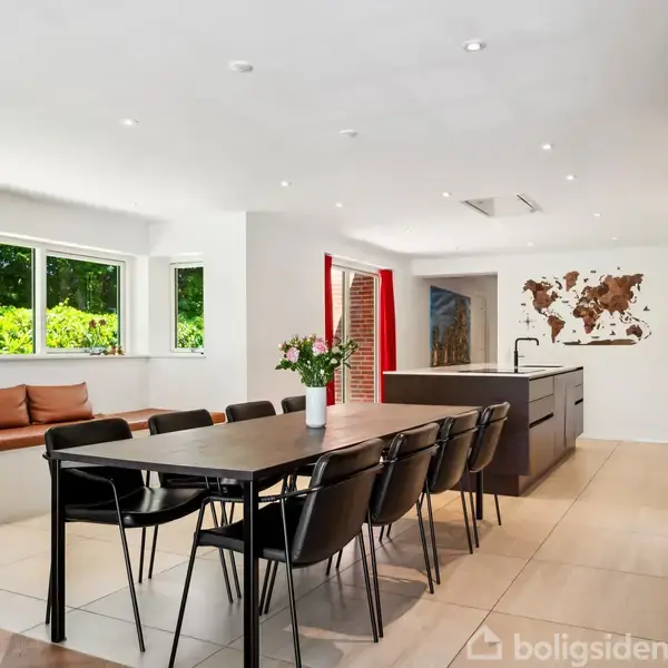 Langbord med sorte stole står i køkkenalrum. Bordet har en vase med blomster. Baghaven ses gennem store vinduer, og der hænger et verdenskort på væggen.