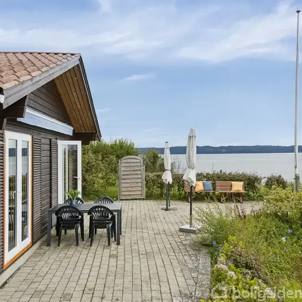 Et træhus med terracotta-tag står ved en brostensbelagt terrasse med sorte stole og en parasol. Omgivet af buske, tilbyder stedet udsigt over en rolig sø i baggrunden.