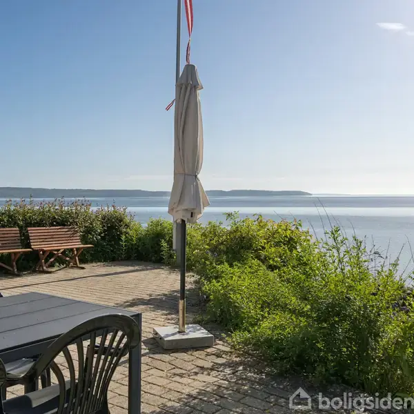 Et bord med stole står på en terrasse med udsigt til havet. To lukkede parasoller står ved siden af bænken, omgivet af buske og klart himmel.