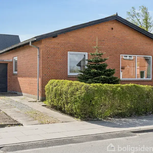 En étplans murstensvilla med et sort tag står frit. En garage ses til venstre. Foran huset er der en lille busk og en fortov.