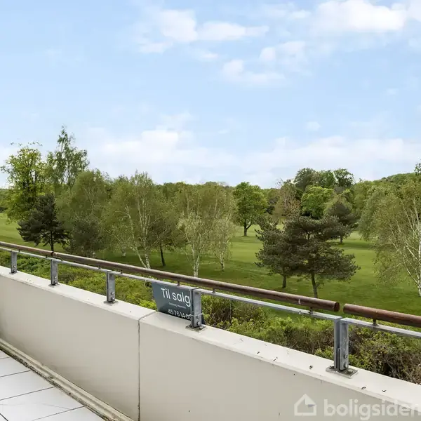Terrasse med flisebelægning og potteplanter vender mod en frodig grøn park med spredte træer. Skilt med teksten "Til salg" er placeret på rækværket. Himmel er klar.