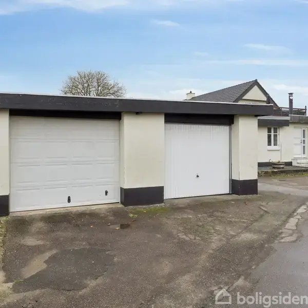 Garage med to hvide porte står på asfaltvej ved siden af et hvidt hus med skråtag og store vinduer; omgivet af grønne buske under en blå himmel.