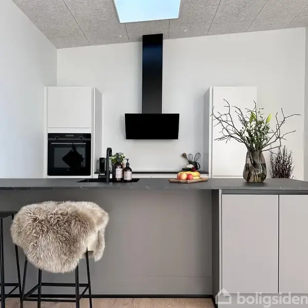 Et moderne køkken med en central ø dækket af grå bordplader. En højstol med fåreskind står ved øen. Baggrunden har indbygget ovn, køkkenredskaber og en vase med grene.