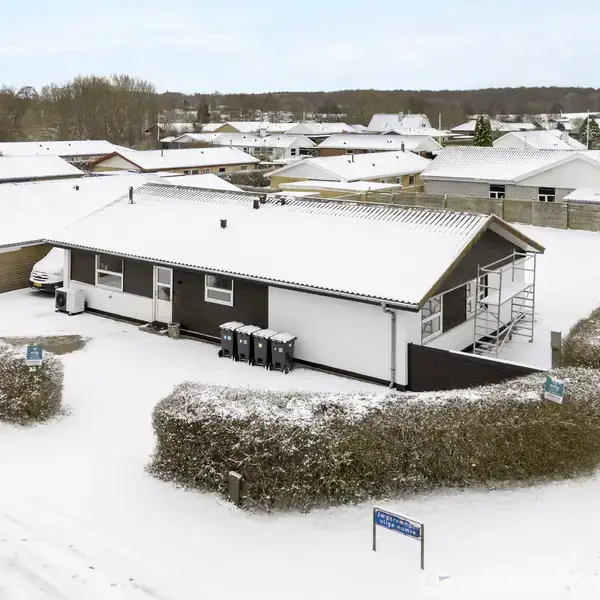 Et hus med fladt tag står stille i et snedækket boligområde. Hække omgiver huset, og flere lignende huse ses i baggrunden. Skilt på sneen: "Gunnar Christensens vej".