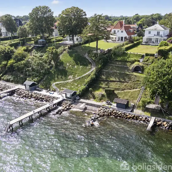 Strandvillaer ligger idyllisk på en skråning, omgivet af grønne haver og træer. Flere små bådebroer strækker sig ud over det klare vand ved kysten, der afslører en rolig, malerisk udsigt.