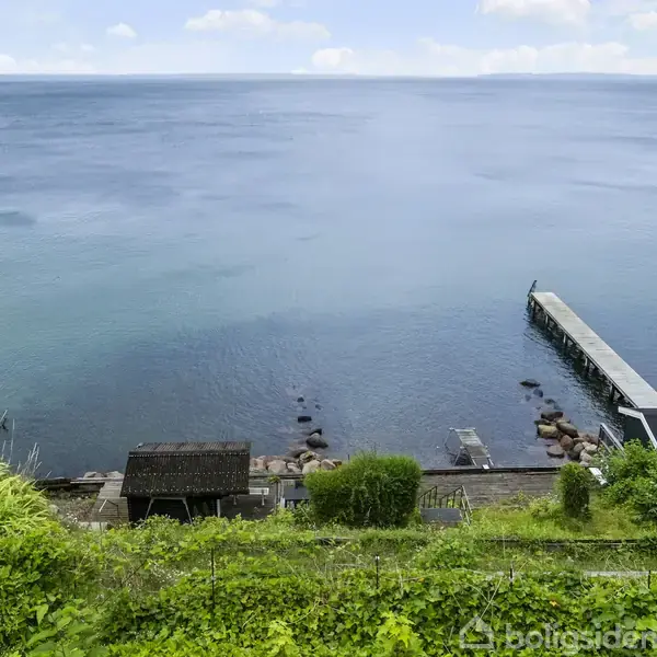 En lang mole strækker sig ud i havet fra kysten. Omgivet af frodige grønne planter og en træbeklædt bakke i forgrunden, med blåt hav vuggende stille i baggrunden.