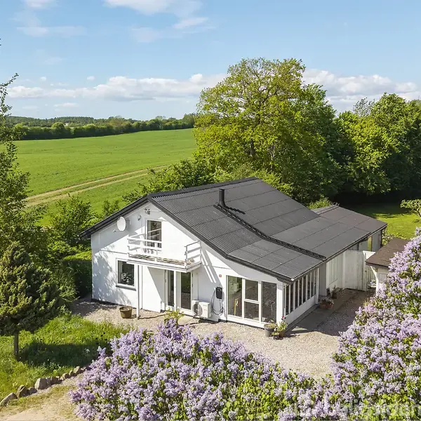 Et hvidt hus med mørkt tag står i en landskabelig have omringet af frodige træer og blomstrende buske. I baggrunden ses åbne marker under en blå himmel.