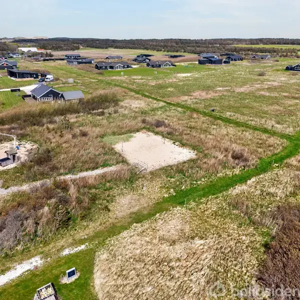 Græs- og buskområde med plan jordstykke i midten. Legestativ til venstre. Omgivet af huse og åbne marker under en klar himmel.