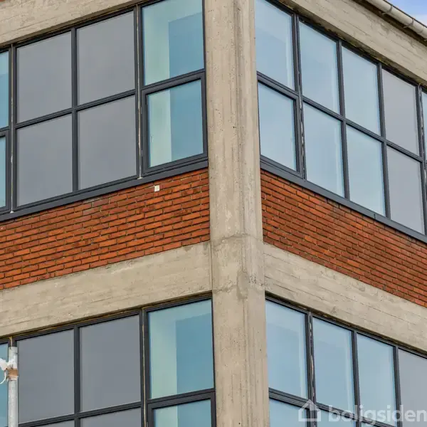 Bygning med store vinduer og murstensvægge. Betonsøjler indrammer glasset, og facaden er moderne. Sollys reflekteres svagt i glasset. Ingen tekst bortset fra vandmærket "Boligsiden".