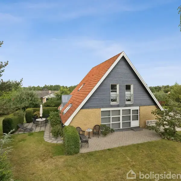 Et A-formet hus med rødt tag og grå facade står i en velplejet have. Rundt om huset er der buske og træer. En terrasse med havemøbler ses til venstre.