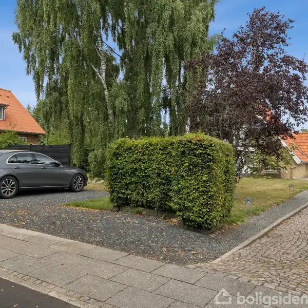 En sølvfarvet bil parkerer på en grusvej foran et hus med rødt tegltag. Omkring bilen er der hække, træer og et brolagt fortov.