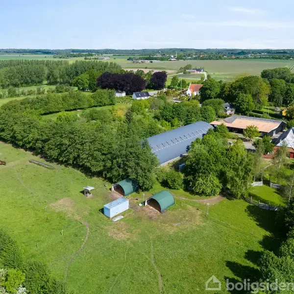 En stor grøn mark med træer omgiver flere landbrugsbygninger, herunder drivhuse og staldlignende strukturer. Nogle få huse er synlige i baggrunden, omgivet af tætte skovområder.