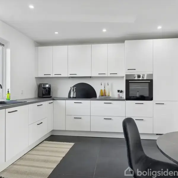 Hvidt køkken med moderne skabe og skuffer, indeholder en integreret ovn og kaffemaskine; ligger i solbeskinnet rum med stort vindue. Rundt spisebord med sorte stole står til højre.