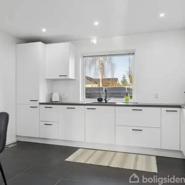 Hvidt køkken med moderne indretning, hvor skuffer og skabe er i brug. Vindue i baggrunden giver udsigt til solrig have. Gråt tæppe ligger på mørkt flisegulv.