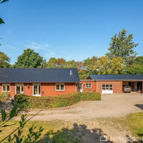 Rødt murstenshus med sorte tagsten står i en lysning omgivet af buske og træer. Foran huset er en grusvej, der fører til to garager. Klar, blå himmel ovenfor.