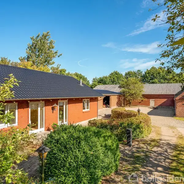 Gårdhus står stille omkranset af træer, med tilhørende udhuse og en gårdsplads. Landlig mark og skovområde ses i baggrunden under en klar blå himmel.