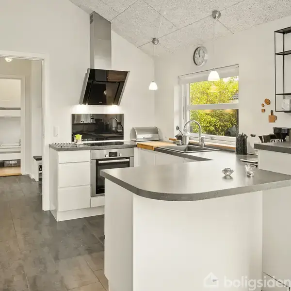 Et moderne køkken med hvide skabe og grå bordplader, hvor en kogeplade og emhætte er centralt placeret. Store vinduer lysner rummet, og der er åben adgang til en gang.