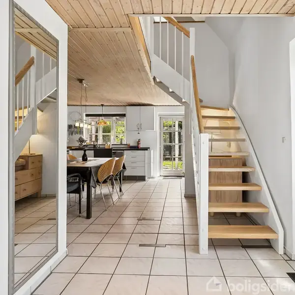 Trappe af træ fører til første sal i et moderne, lyst rum. Spisebord med stole står til venstre i en flisebelagt gang med store spejle og synlige trælofter.