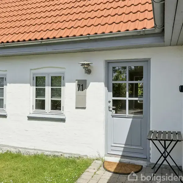 Hvidt hus med rødt tegltag. En grå dør ved siden af to vinduer. Nummeret "71" hænger ved døren. Græsplæne og flisegang i forhaven.