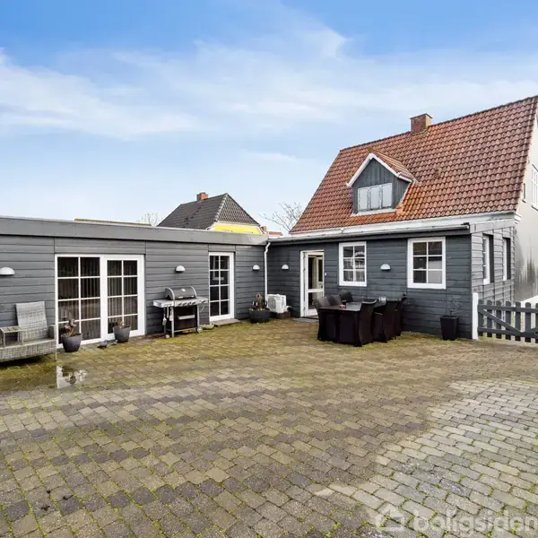 Et gråt hus med rødt tag står stille i en brolagt gårdhave. Havemøbler og griller er placeret ved siden af huset, omkranset af et lavt stakit.