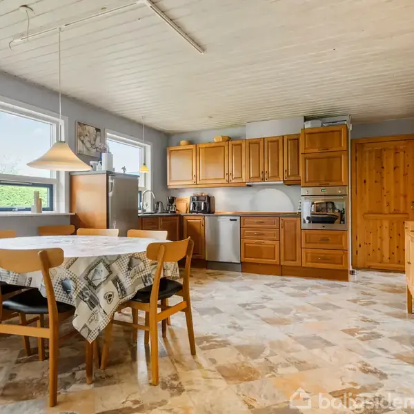 Et rummeligt køkken med spisebord i midten dækket af en mønstret dug. Træstole omkranser bordet. Skabe i træ, vinduer mod venstre, og køkkenapparater fylder væggene.