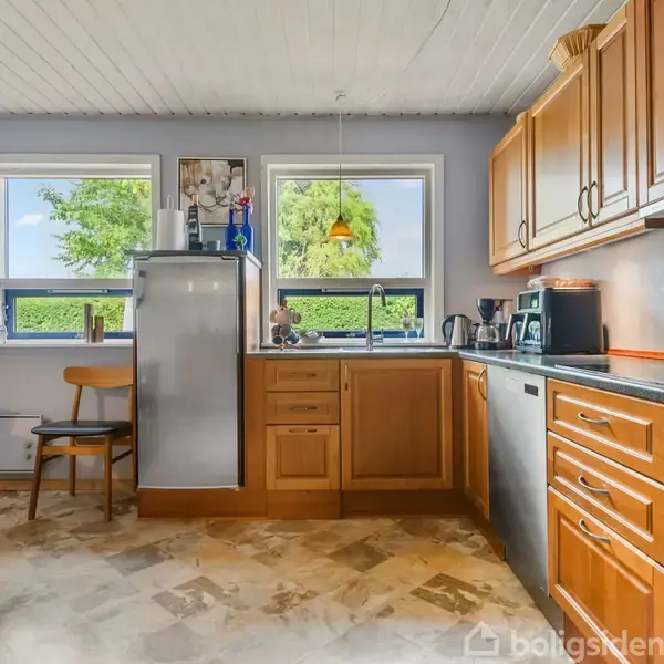 Et køkken med træskabe og sorte bordplader indeholder en grå opvaskemaskine, kaffemaskine, og forskellige køkkenapparater. Spisebord med stole står ved siden af tre vinduer, der slipper lys ind.