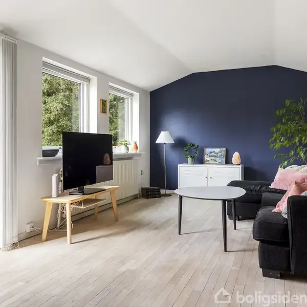 Stue med sort sofa og sofabord foran en blå væg. Fladskærms-TV på træbord. Store vinduer og åben dør til terrasse. Gulvlampe og planter tilføjer dekoration.
